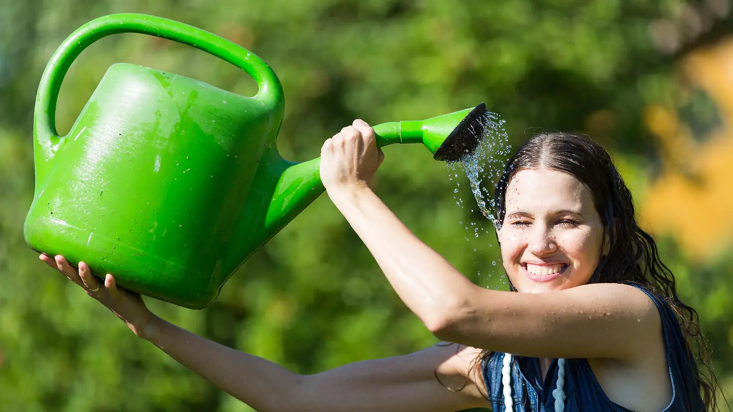 Eine lächelnde Frau gießt sich mit einer Gießkanne Wasser über den Kopf.