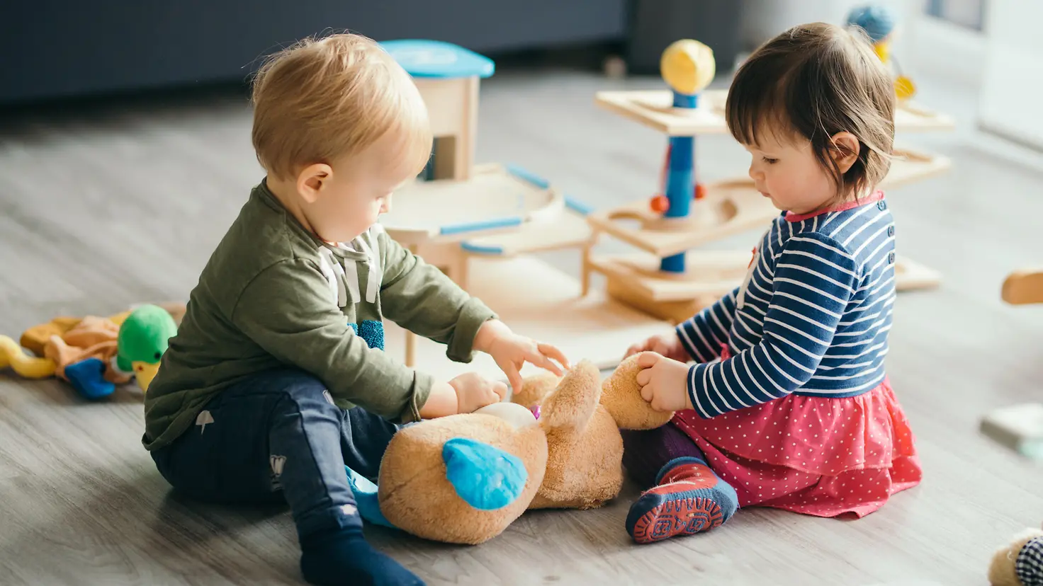 Zwei Kleinkinder sitzen auf dem Boden und spielen mit einem Teddybär.