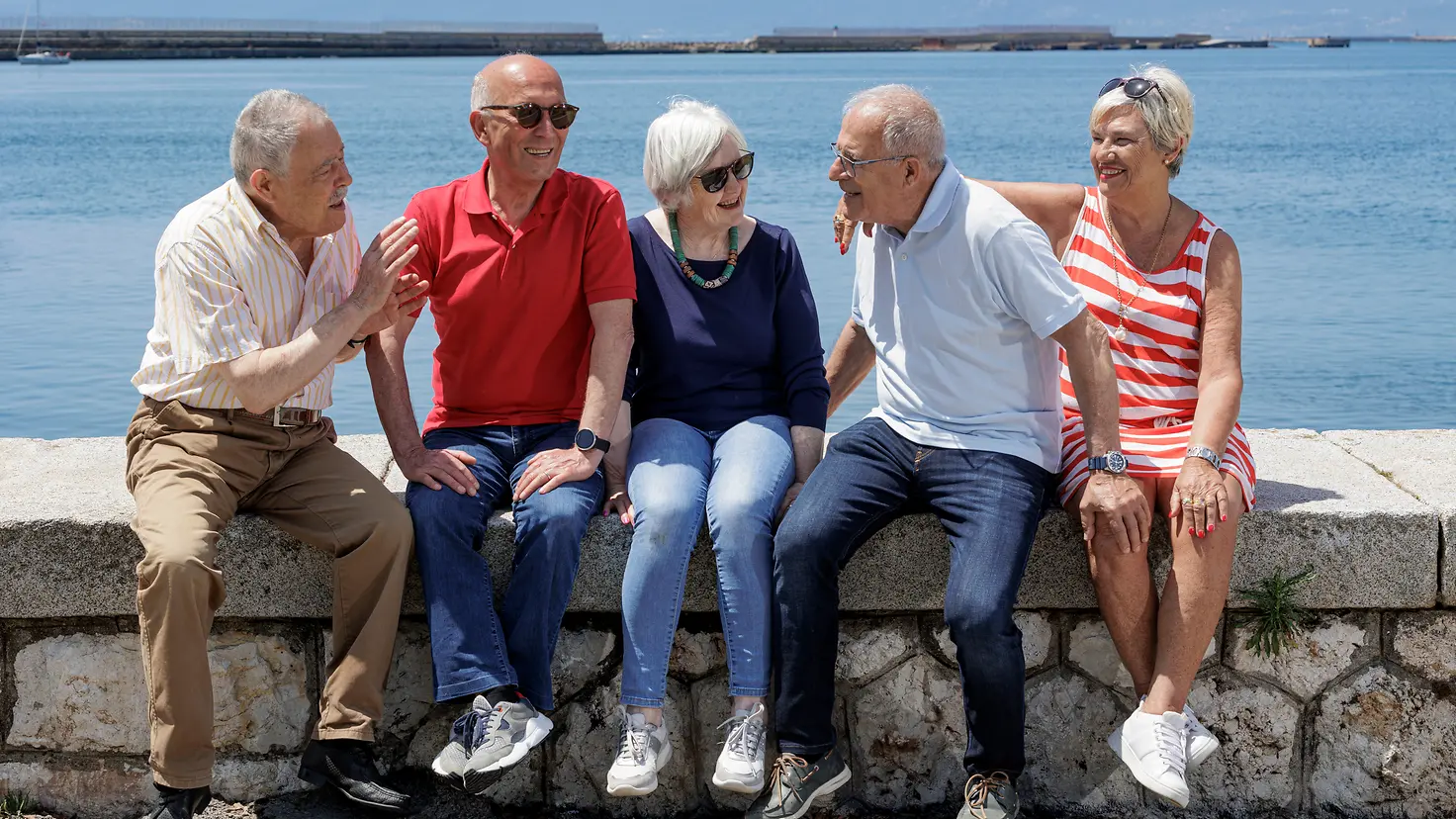 Zu sehen sind fünf ältere Personen, die auf einer Mauer am Meer sitzen