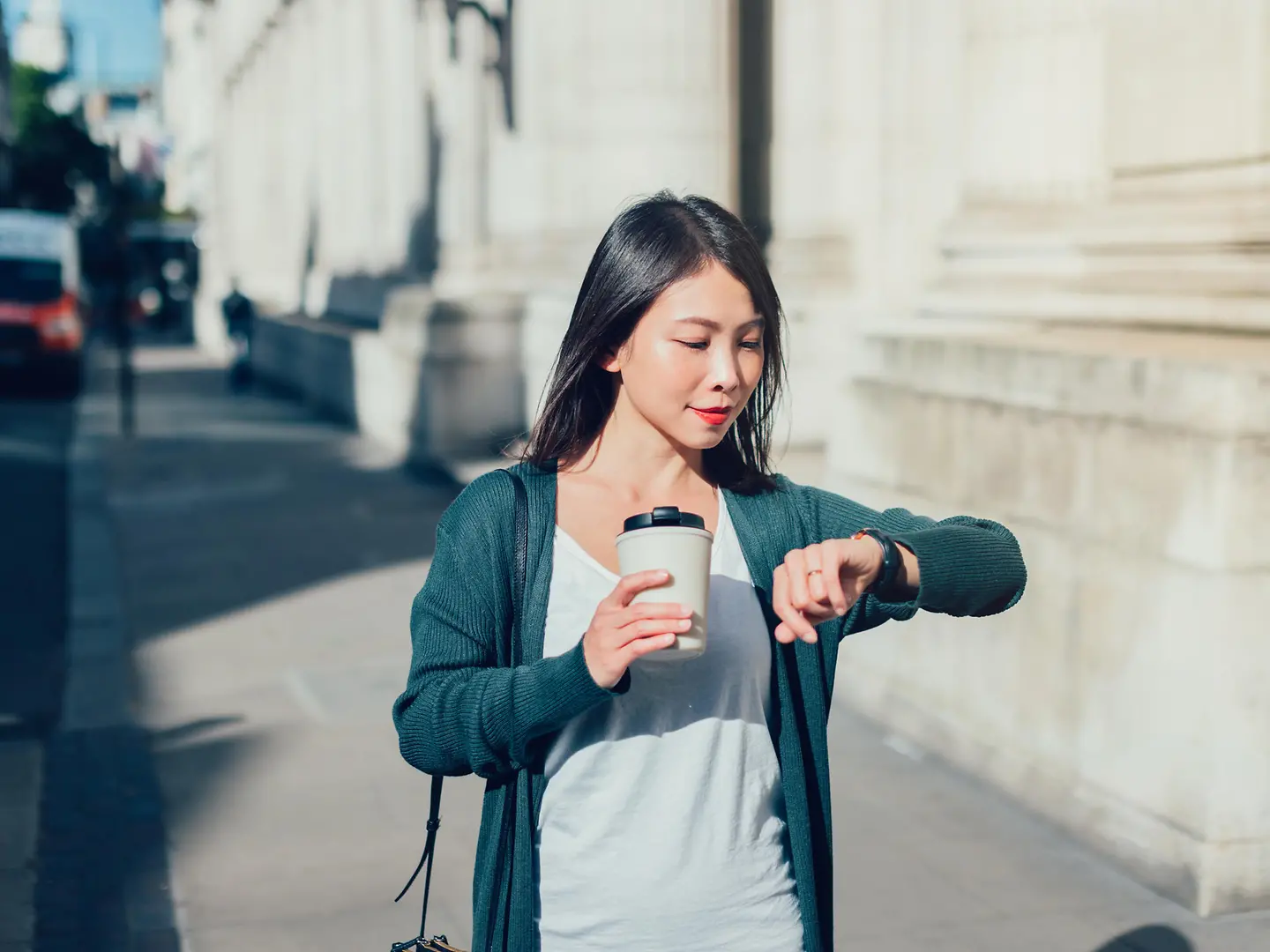 Eine junge Frau mit Kaffeebecher in der Hand schaut auf die Uhr. 