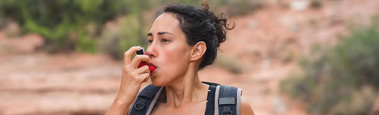 Eine Frau mit Rucksack benutzt während einer Wanderung ihr Asthmaspray.