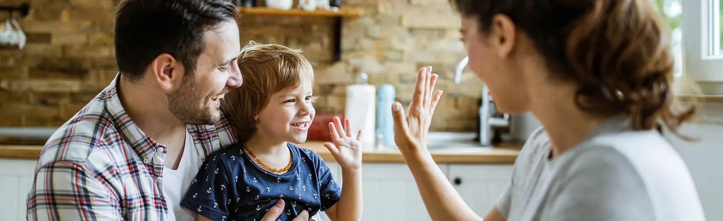 Ein Kind sitzt bei seinem Vater auf dem Arm und gibt seiner Mutter High Five. 