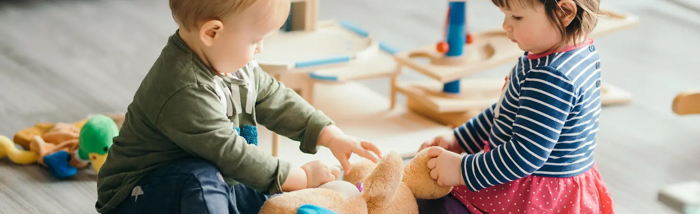 Zwei Kleinkinder sitzen auf dem Boden und spielen mit einem Teddybär.