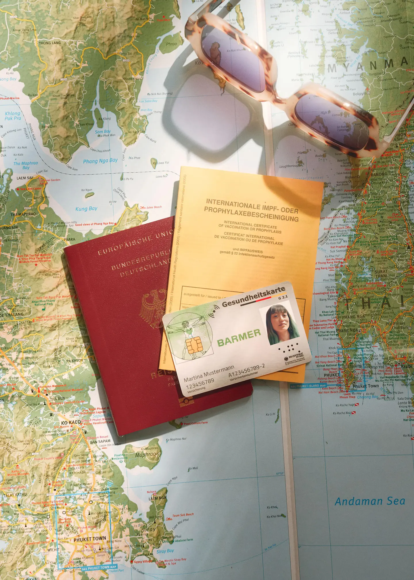 Auf einer ausgefalteten Weltkarte liegen eine Barmer-Versichertenkarte, ein Reisepass, ein Impfausweis und eine Sonnenbrille. 