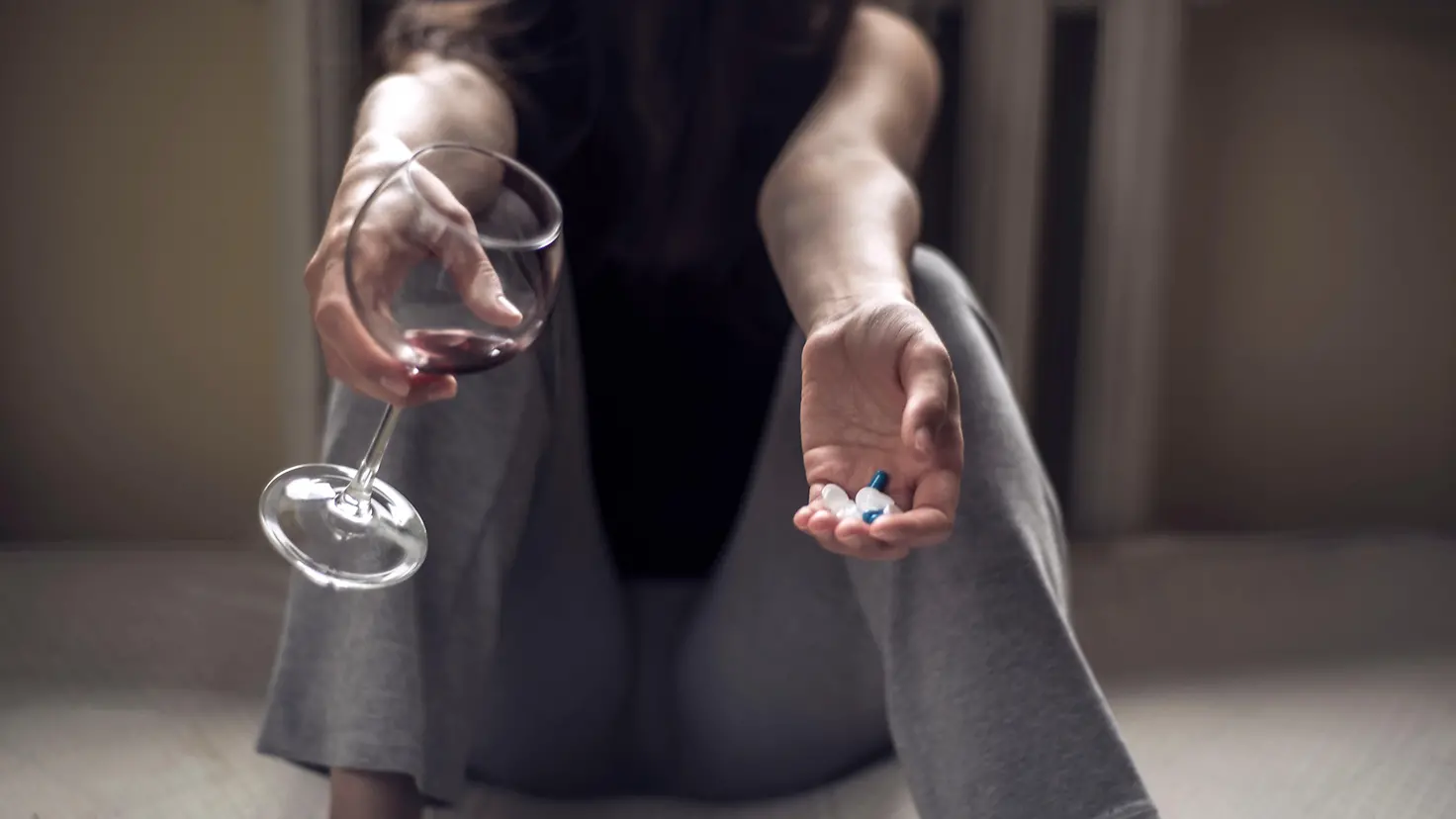 Zu sehen ist eine sitzende Frau, mit einem Rotweinglas in der einen und Pillen in der anderen Hand