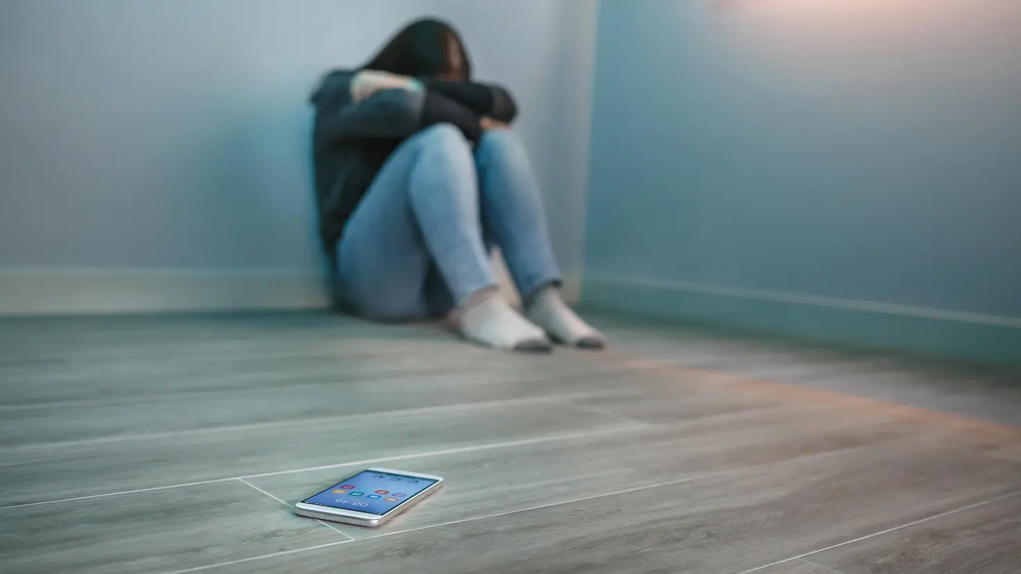 Zu sehen ist eine auf dem Boden sitzende junge Frau mit verschränkten Armen vor dem Gesicht und ein Smartphone