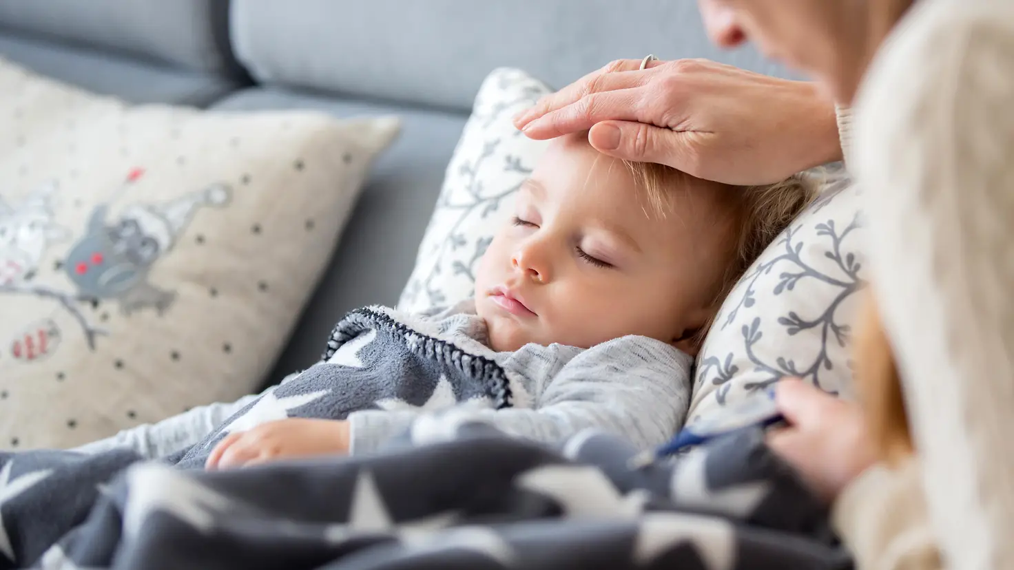 Zu sehen auf diesem Bild ist ein auf der Couch liegendes Kind mit geschlossenen Augen. Die Hand der Mutter liegt auf der Stirn des Kindes.