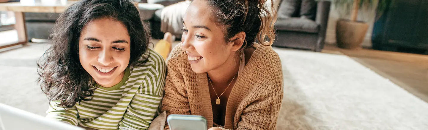 Two young women have made themselves comfortable on the carpet with their smartphone and tablet.