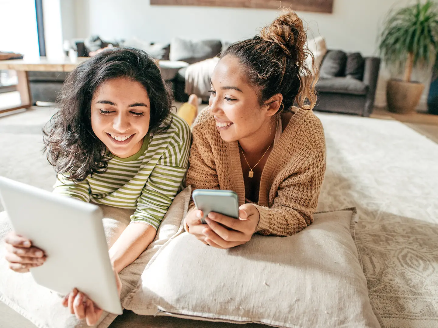 Two young women have made themselves comfortable on the carpet with their smartphone and tablet.