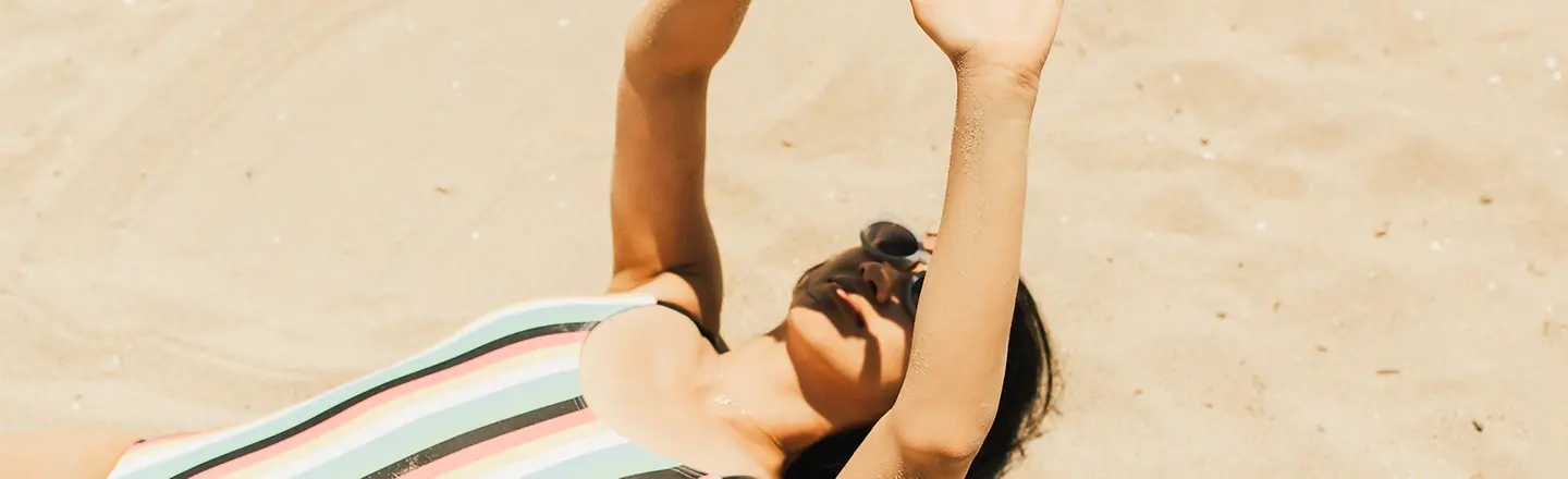 Junge Frau liegt im Sand und hält sich schützend die Hände vors Gesicht. 