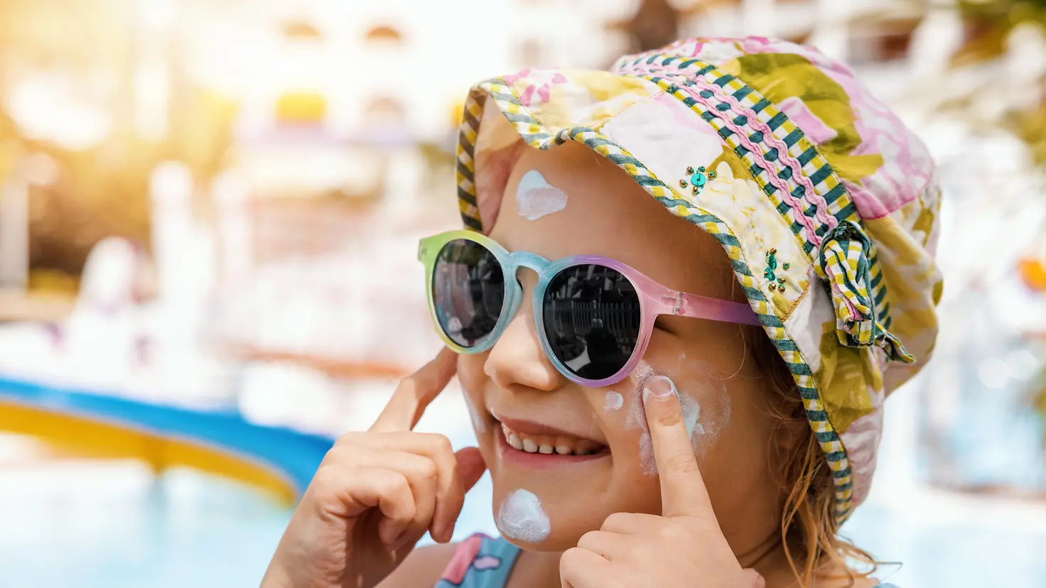 Zu sehen auf diesem Bild ist ein kleines Mädchen mit Sonnenhut und Sonnenbrille, die mit ihren Zeigefinger Sonnencreme in ihrem Gesicht verteilt und dabei lacht.
