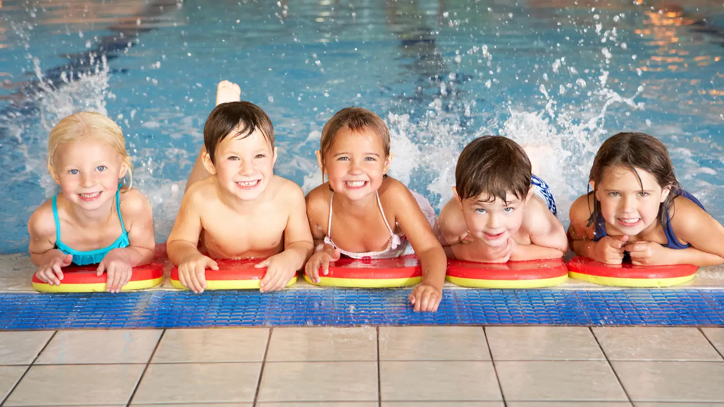 Zu sehen auf diesem Bild sind fünf Kinder, die sich am Rand eines Schwimmbeckens im Wasser aufhalten. Sie stützen sich auf ihren Schwimmhilfen ab und planschen mit dem Beinen.