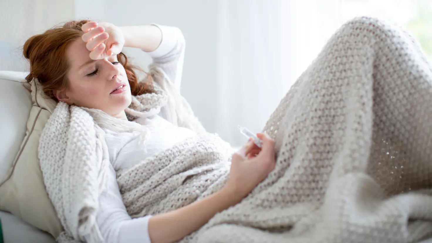 Zu sehen ist eine Frau unter einer wärmenden Decke mit Fieberthermometer in der Hand
