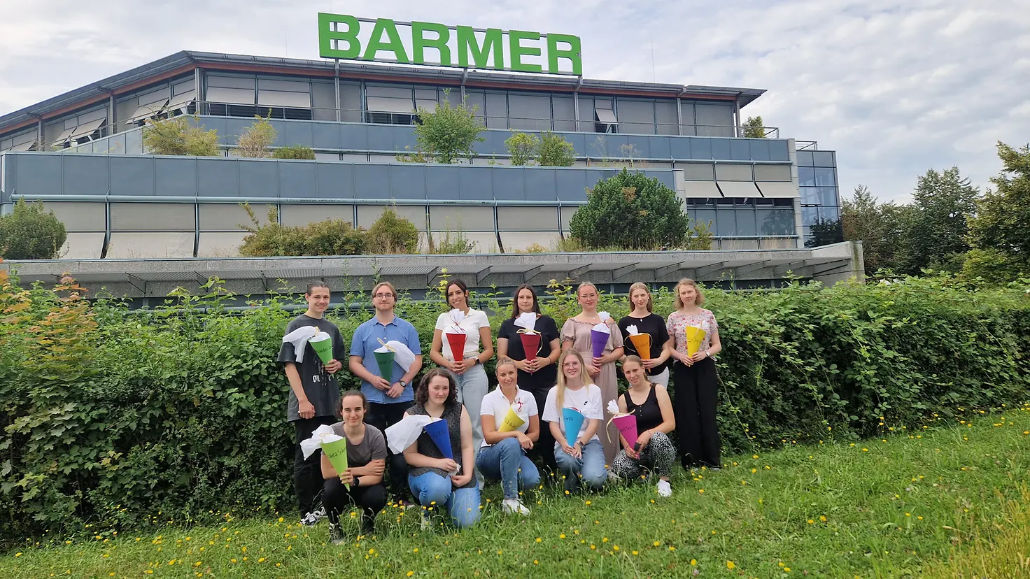 Zwölf junge Frauen und Männer mit Schultüten in der Hand stehen und hocken vor einem Bürogebäude mit der Aufrschrift 'BARMER'