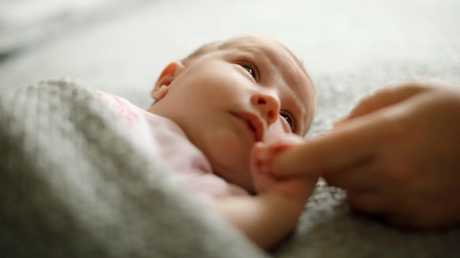 Ein Baby liegt unter einer Decke, greift den Finger einer erwachsenen Person und richtet seinen Blick in deren Richtung