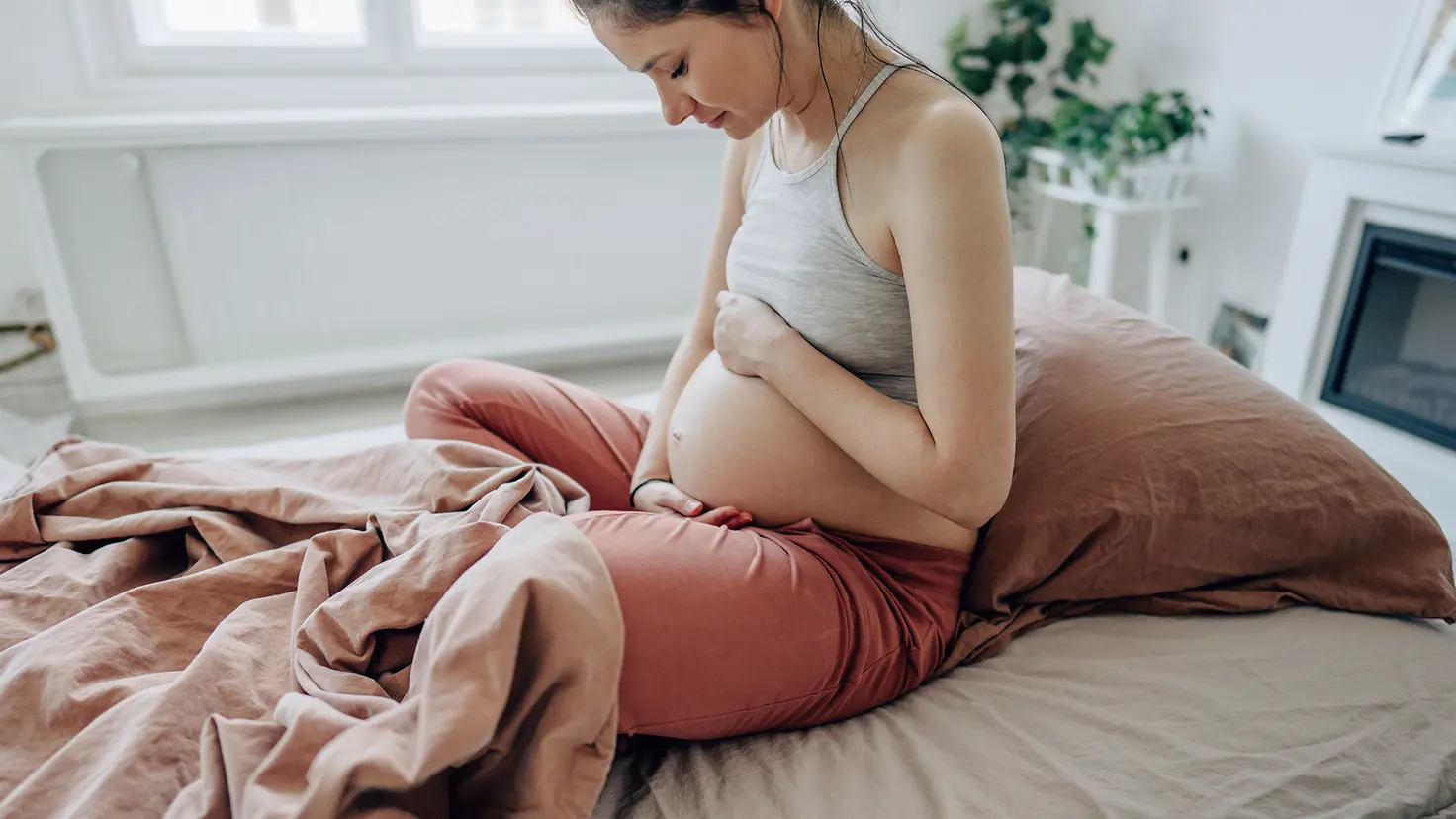 Eine schwangere Frau sitzt auf einem Bett und streichelt ihren Bauch