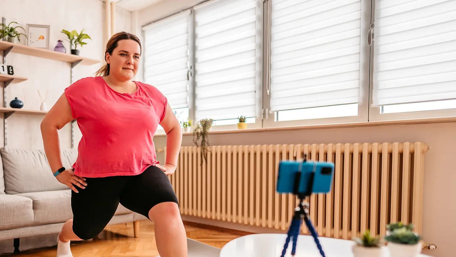 Junge übergewichige Frau macht zu Hause Sport und nutzt dazu ihr Handy als Monitor für die Übungen