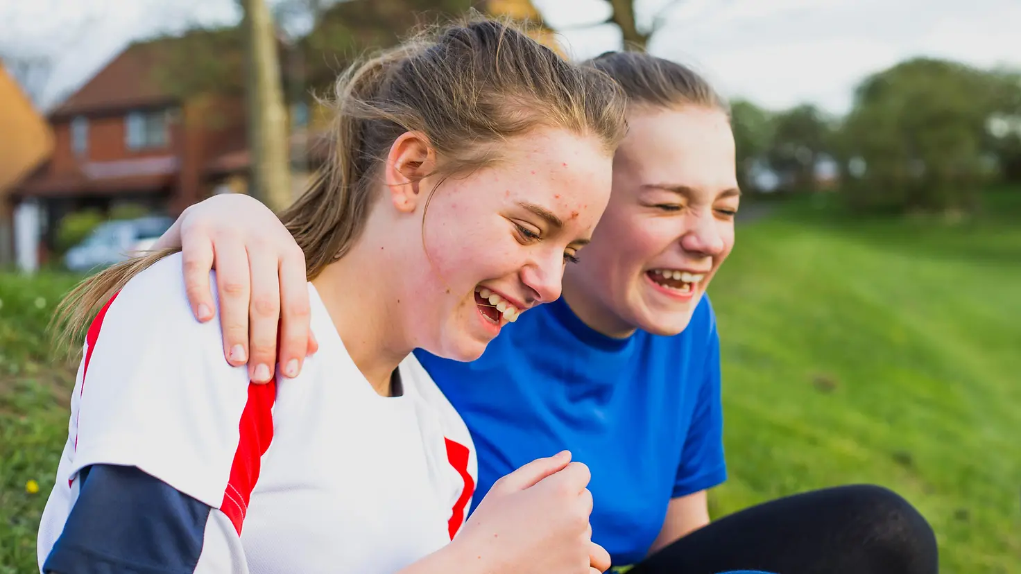 Zwei Mädchen im Teenageralter sitzen nebeneinander im Gras und lachen