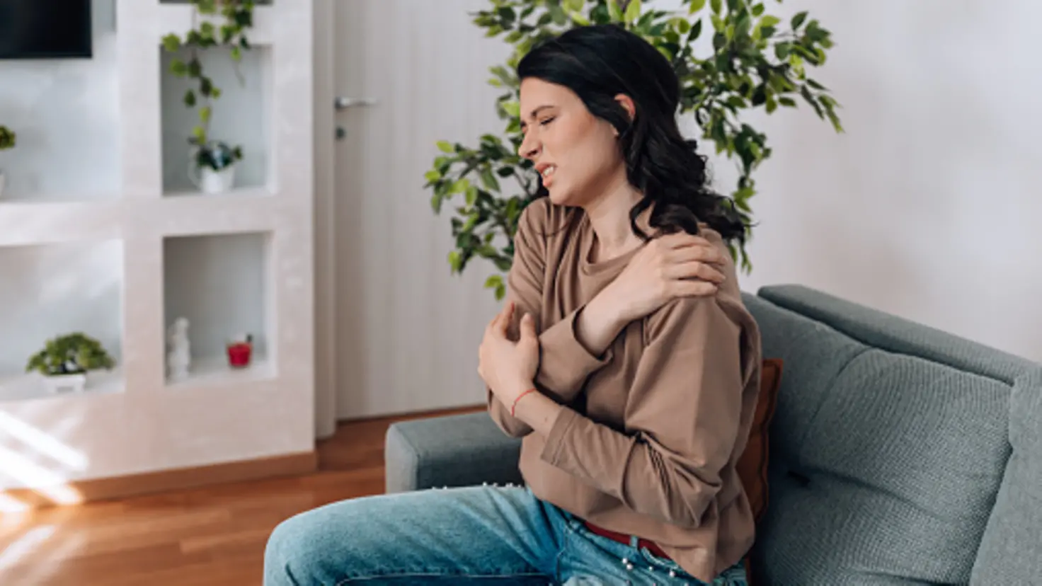 Eine junge Frau sitzt auf einem Sofa und hält sich mit schmerzverzerrtem Gesicht die Schulter. Sie wirkt angespannt und scheint unter Schmerzen zu leiden. Im Hintergrund sind Zimmerpflanzen und ein modernes Regal zu sehen.