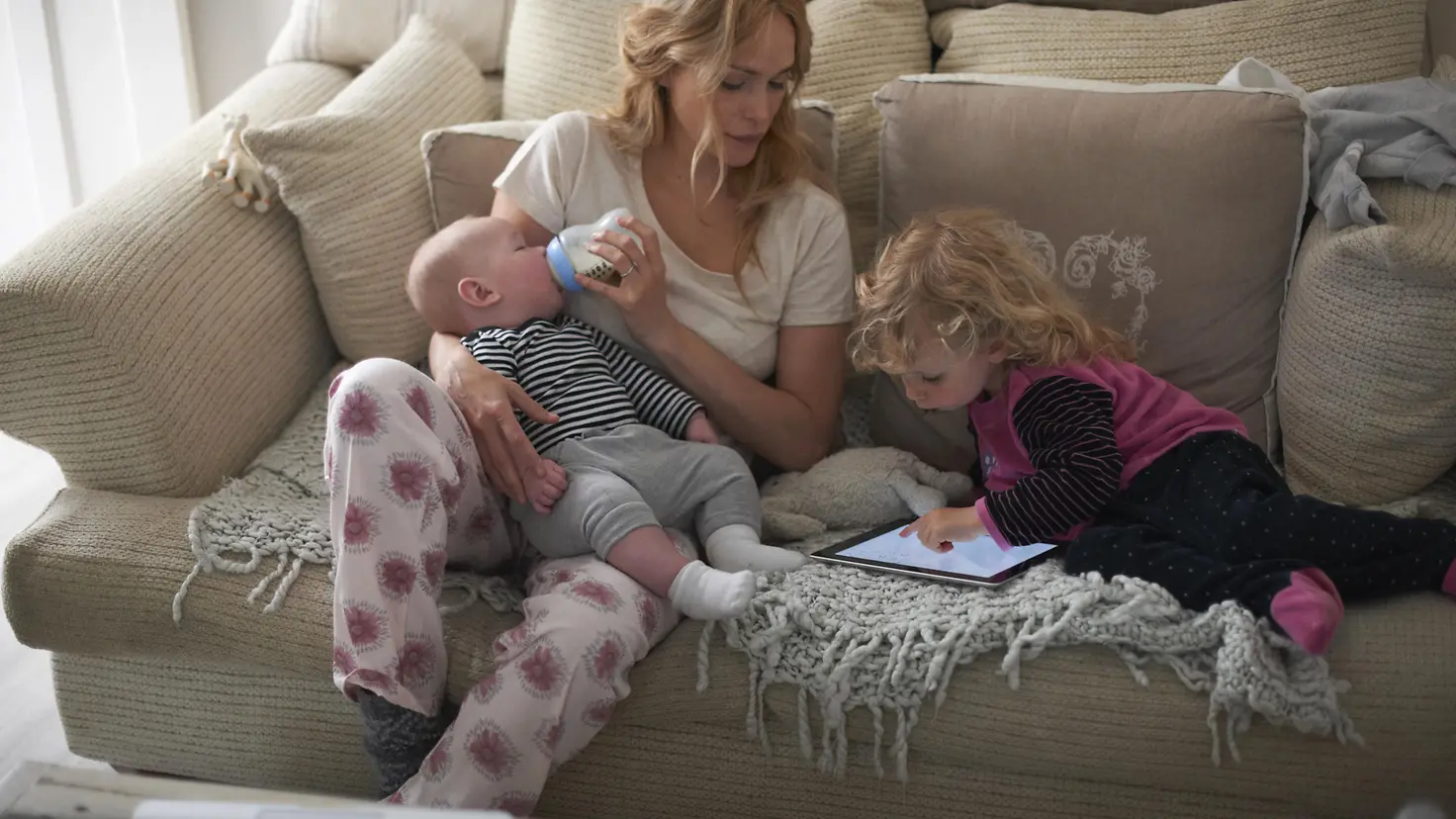 Eine Mutter sitzt im Wohnzimmer auf einem Sofa und füttert ihr Baby mit einer Flasche, während ihr kleines Kind neben ihr auf einem Tablet spielt.