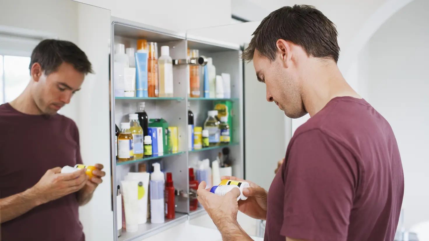 Mann steht im Bad vorm geöffneten Spiegelschrank und hält zwei Dosen Tabletten in der Hand und betrachtet diese