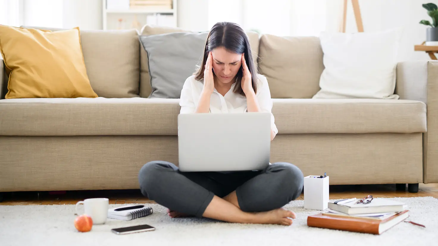 Frau mit Spannungskopfschmerzen sitzt mit Laptop auf dem Wohnzimmerfußboden.