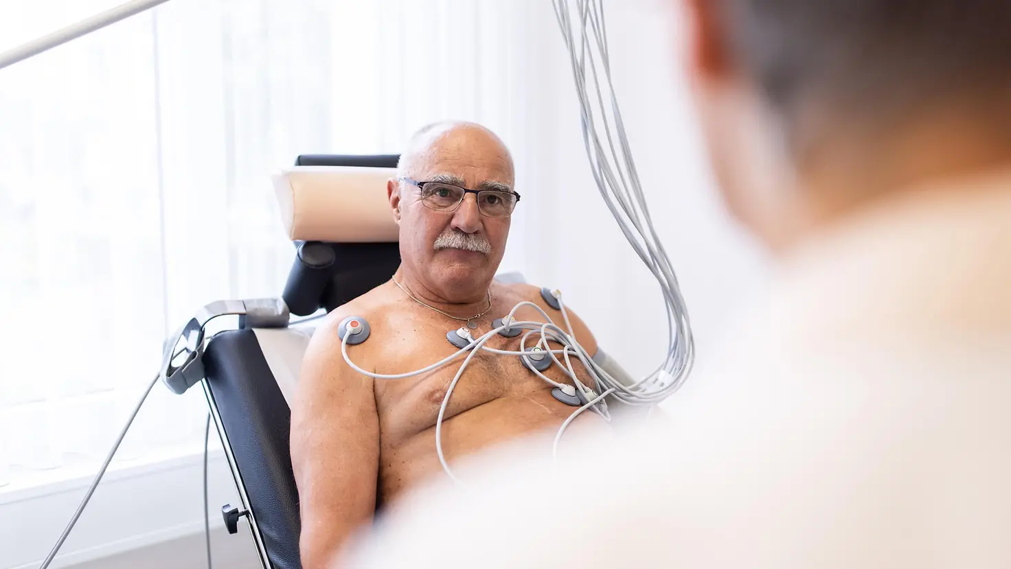 Älterer männlicher Patient mit EKG-Sensor auf der Brust schaut einen Arzt in der Klinik an. Der Senior führt einen Herzbelastungstest in der kardiologischen Klinik durch.