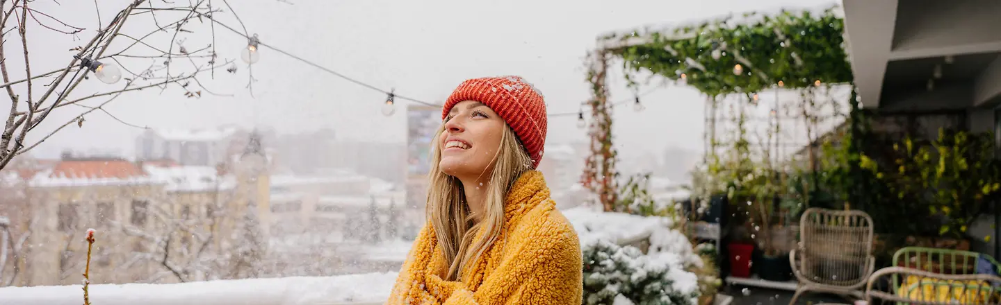 Eine junge Frau steht im Schnee auf einem Balkon in eine Decke eingewickelt