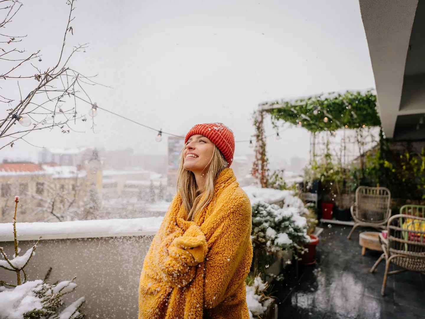 Eine junge Frau steht im Schnee auf einem Balkon in eine Decke eingewickelt