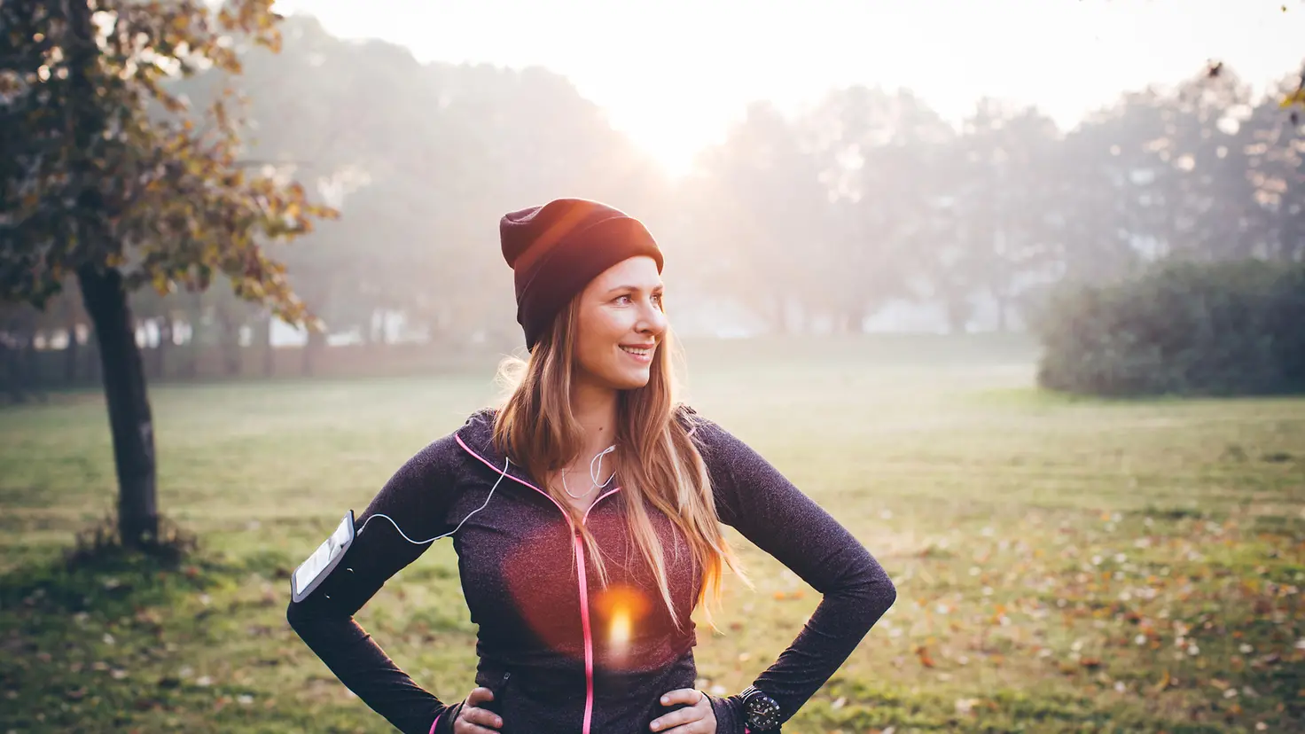 Junge Frau in Sportklamotten steht lächelnd im Park.