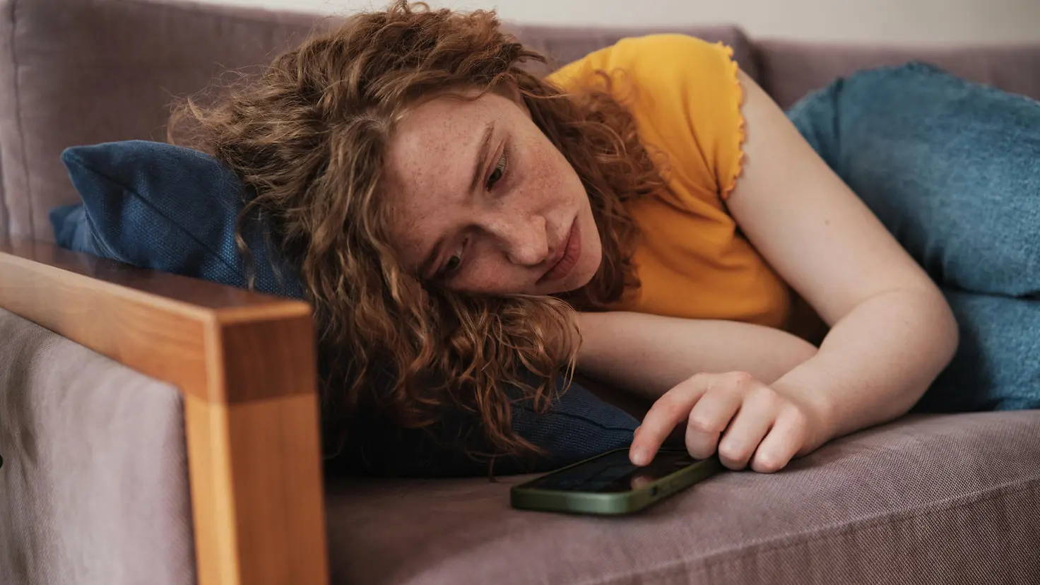 Junge Frau mit roten Haaren liegt auf einer Couch und schaut auf ihr Handy