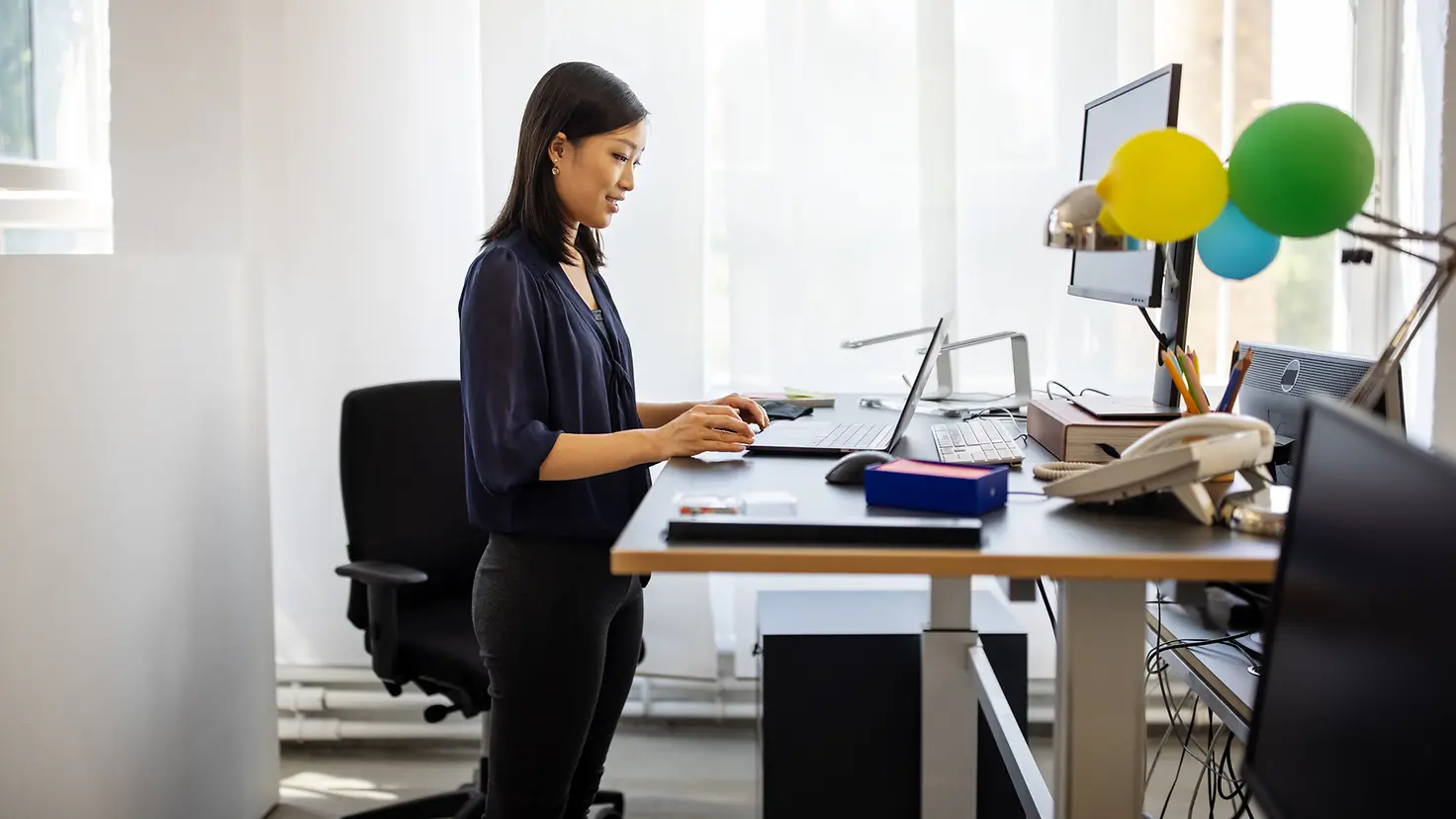 Eine Frau arbeitet im Stehen an ihrem Schreibtisch im Büro, um Beschwerden durch einen Bandscheibenvorfall zu lindern.