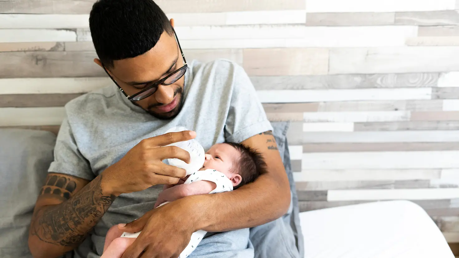 Junger Vater sitzt auf dem Bett und gibt seinem Baby das Fläschchen